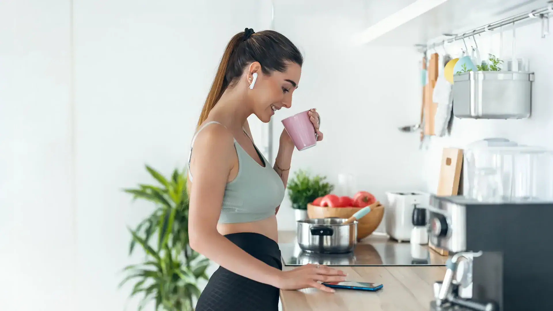 Woman in a sports bra and earphones enjoys a cup of coffee, showcasing a blend of fitness and relaxation.