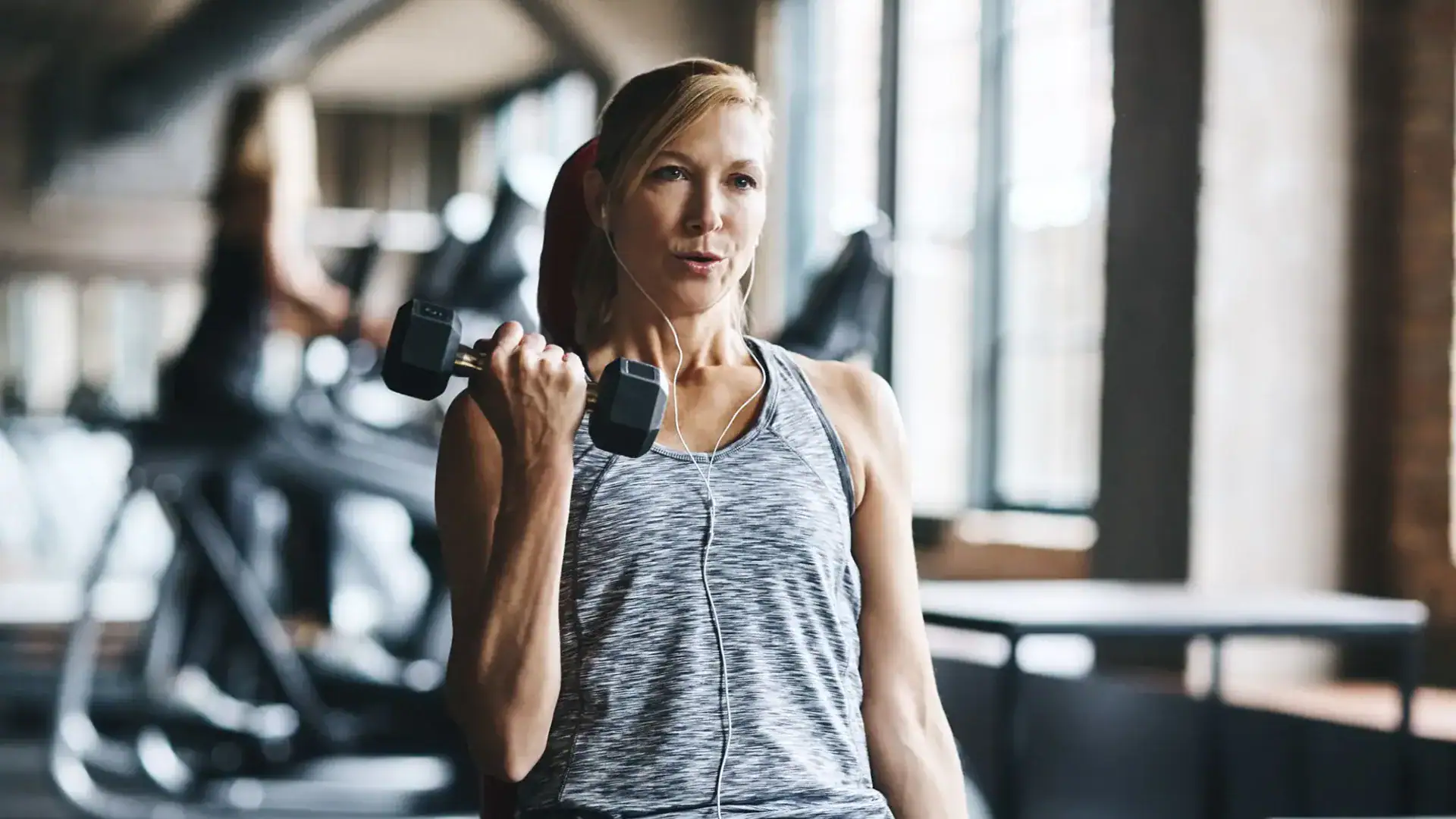 A woman in athletic wear lifts a dumbbell with one arm while listening to earphones in a gym with large windows.