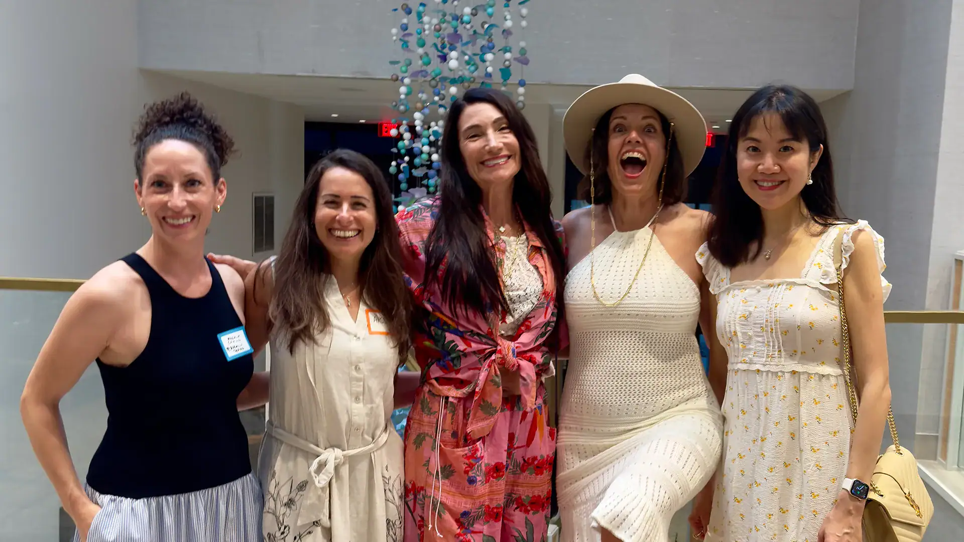 Five women smile and pose together at a networking event inside a bright, modern venue.
