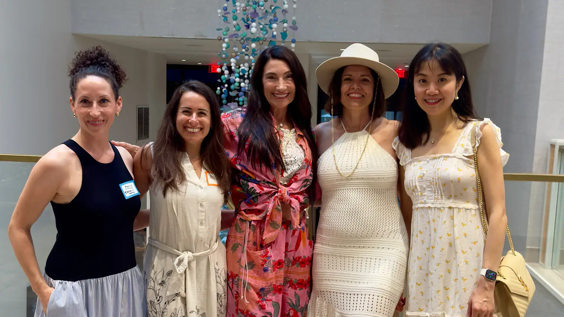 Five women smile and pose together at a networking event inside a bright, modern venue.