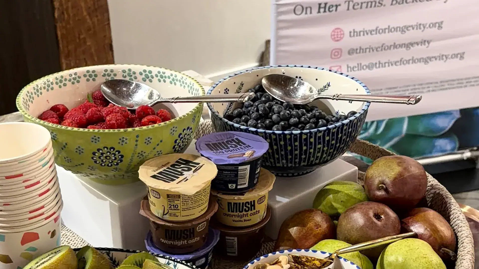 A healthy refreshment station features bowls of fresh raspberries and blueberries, pears, kiwi, and protein-rich overnight oats arranged for attendees.