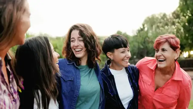 Five women laughing together in a sunny park, enjoying each other's company and the outdoors.