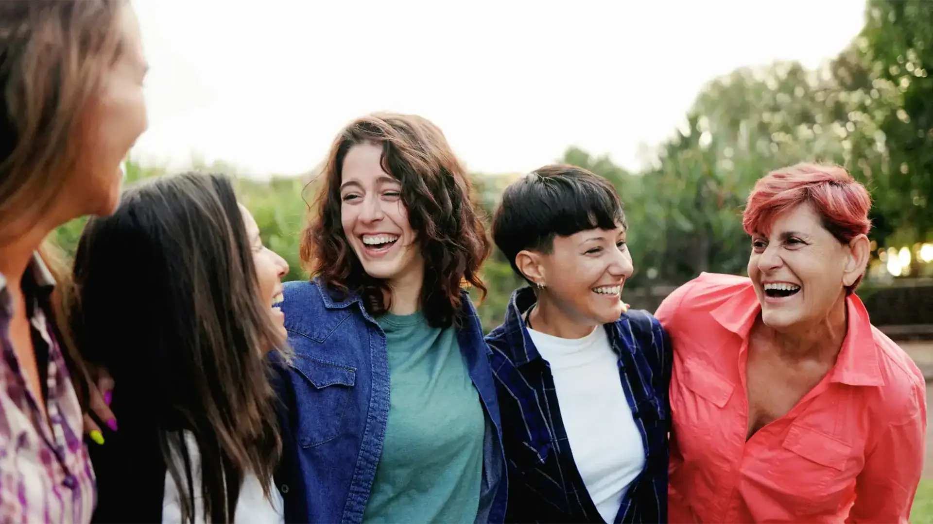 Five women laughing together in a sunny park, enjoying each other's company and the outdoors.