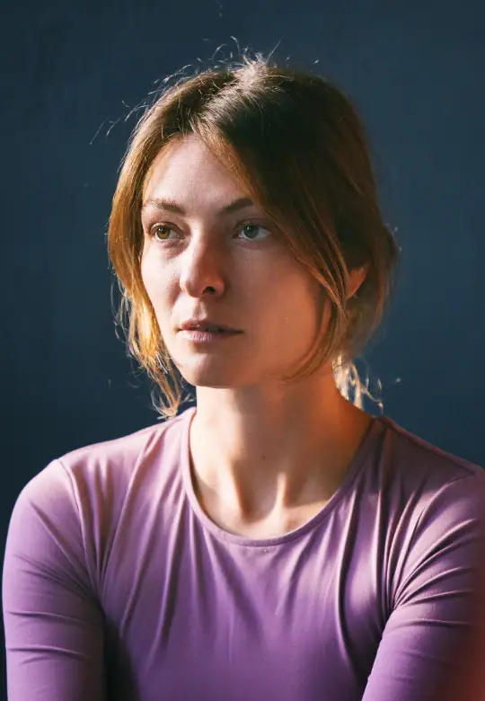 A woman in a lavender top gazes thoughtfully into the distance, her expression calm and reflective against a softly lit dark background.
