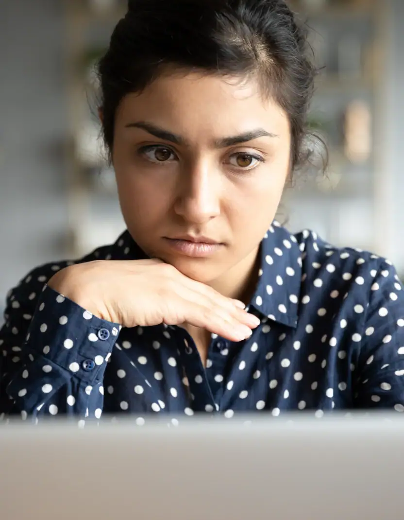 A woman in a polka-dot blouse rests her chin on her hand while looking thoughtfully at a screen, conveying concentration, reflection, or concern.