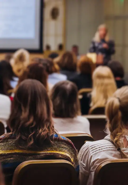 Audience members seated in a conference room listen to a speaker presenting at the front.
