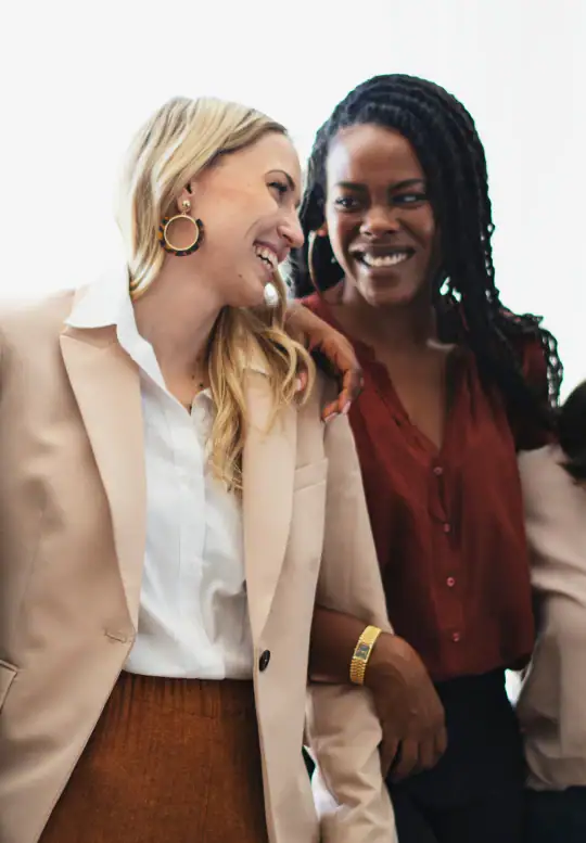 Two women of different backgrounds share a joyful moment together, smiling and laughing, representing friendship, support, and connection among women.