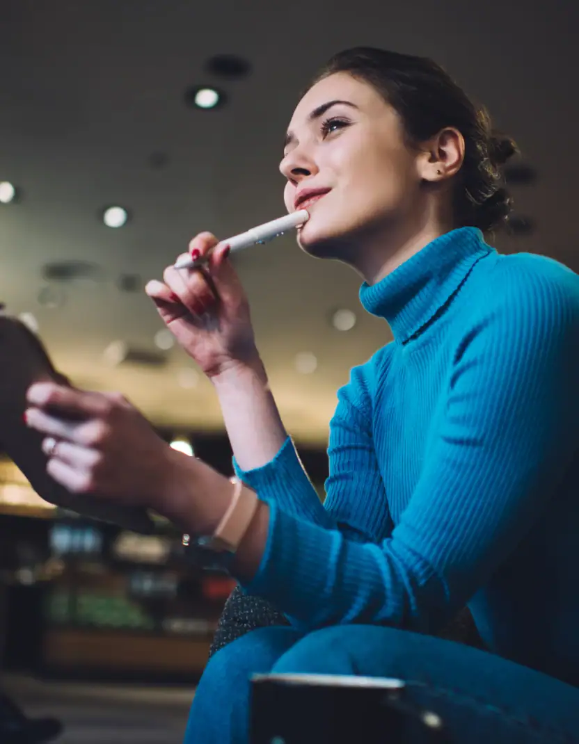 A woman in a blue turtleneck sits thoughtfully with a tablet and stylus in hand, appearing engaged in learning, planning, or taking notes in a modern indoor setting.