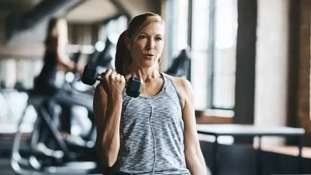 A woman in athletic wear lifts a dumbbell with one arm while listening to earphones in a gym with large windows.
