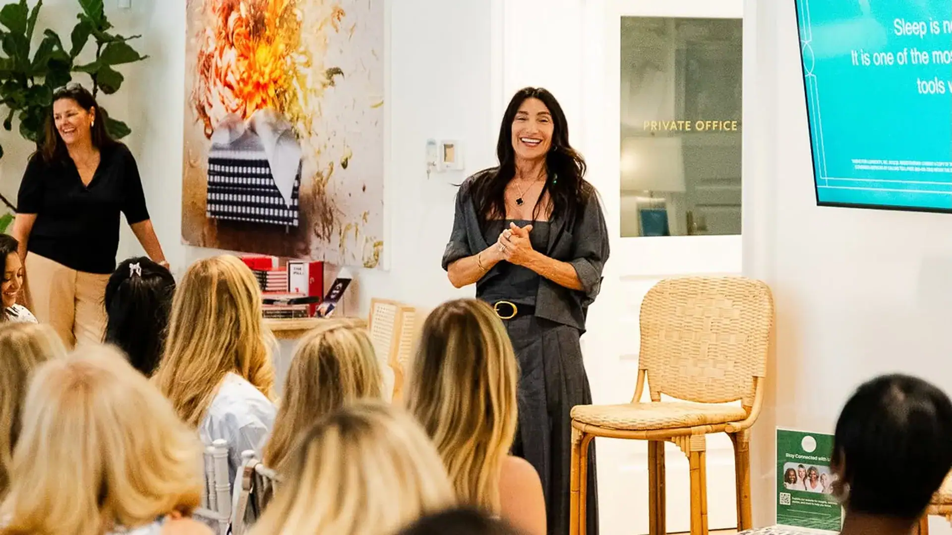 A woman stands at the front of a wellness seminar, smiling as she speaks to an audience seated in rows.