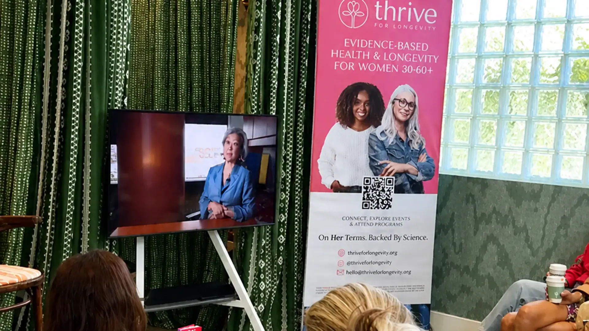 Attendees watch a presentation displayed on a large monitor beside a Thrive for Longevity banner promoting evidence-based health and longevity programs for women.