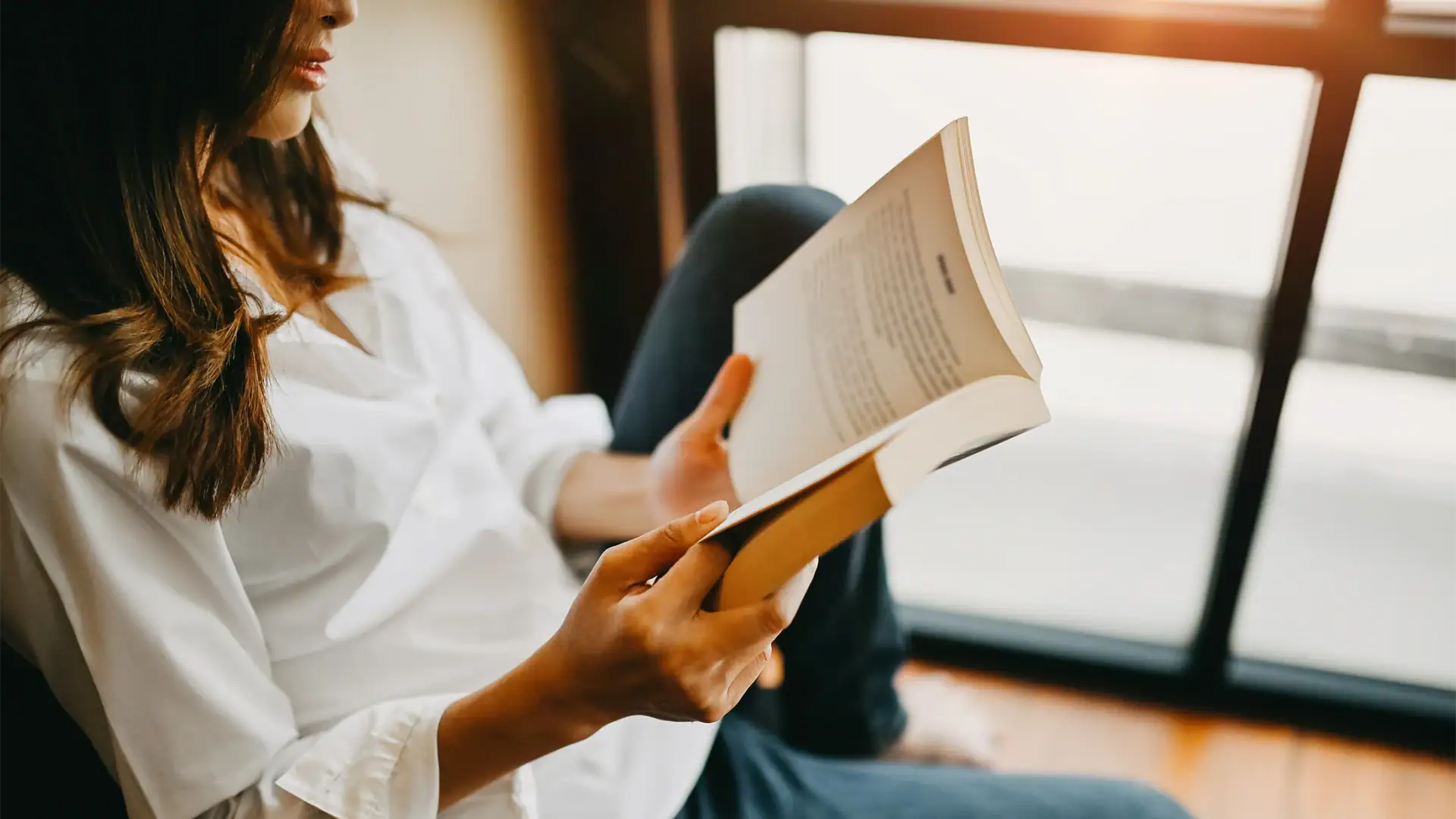 Woman reading a book while sitting by a window, representing lifelong learning, education, and personal wellness.
